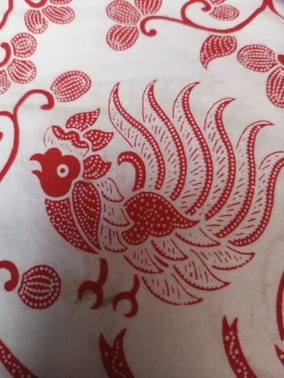 A white fabric features a repeated pattern in red. Stylized flowers with dotted centers and curved vines are scattered across the surface. A central motif depicts a rooster or similar bird with a prominent crest, detailed feathers filled with dots, and small feet. The design style is reminiscent of a hand-printed or batik technique. Minor stains are visible on the fabric near one of the floral patterns.