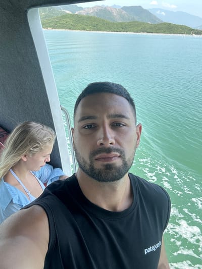 A man is taking a selfie on a boat, with a woman in the background. The man is in the foreground, facing the camera, with a slight beard and short dark hair. He is wearing a black t-shirt with the Patagonia logo on it. The woman is to his left, also on the boat, looking down and away from the camera. She has blonde hair and is wearing a light blue shirt and a white bikini top. The boat is on the water, which is a bright green color, with some white foam visible from the boat's movement. In the background, there are green, tree-covered hills and mountains, with a cloudy sky. The location is likely the coast of Ninh Hoa city, Khanh Hoa province, Vietnam, known for its scenic beauty and coastal activities.