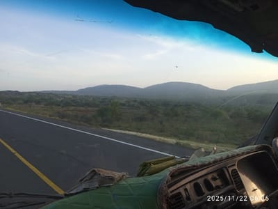 A view from inside a vehicle, looking out onto a highway. The road stretches into the distance, flanked by scrubland and low hills under a hazy sky. The vehicle's dashboard is visible, covered with a green quilted material and personal items. The windshield has visible cracks. A timestamp at the bottom right reads "2025/11/22 06:35".