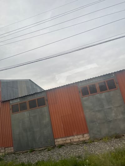 The image depicts a building in Akhuryan, Armenia, characterized by its corrugated metal exterior. The building has sections of orange and gray metal. Windows are visible, framed in a brown material. A gravel and grassy area runs along the base of the building. Power lines are strung overhead. The sky is overcast.