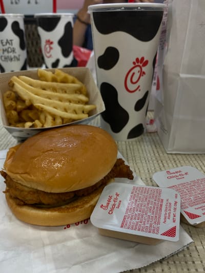This is a close-up, eye-level, medium shot of a Chick-fil-A meal on a table in Hagerstown, Maryland. In the foreground is a chicken sandwich on a bun, with waffle fries in a white paper container behind it. Two Chick-fil-A sauce packets are next to the sandwich. In the background, there are several large Chick-fil-A branded cups with a cow print design. One of the cups has a red straw. The table appears to be a light-colored wood. The overall scene suggests a casual dining experience at a fast-food restaurant.
