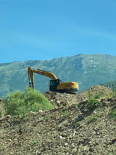 An excavator is shown in a construction site in Prijeradi, Montenegro. The excavator is yellow and is positioned on a pile of dirt. In the background, there is a mountain range. The sky is clear and blue.