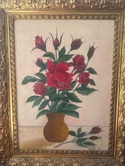 A painting in a gold ornate frame depicts a bouquet of red roses in a brown vase, placed on a table. The background is a light pinkish hue. To the right of the vase, a separate branch with two rosebuds and leaves lies on the table. The artist's signature, "B. Nivelli, 1975," is visible in the bottom right corner. The painting is displayed indoors, and the specific location in Ringsted, Denmark, is not ascertainable from the image.