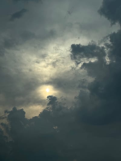 The image is a photograph of a dramatic sky with thick, dark clouds partially obscuring the sun. The sun is visible as a bright, yellow-white orb, radiating light through breaks in the clouds. The clouds are varied in texture and density, with some appearing light and wispy, while others are heavy and ominous. The overall mood is somewhat dramatic and turbulent, suggesting a transition in weather, possibly before or after a storm. The lighting is dynamic, with bright areas where the sun shines through and deep shadows in the denser cloud formations. There is no visible text, people, or man-made objects. The scene is entirely focused on the sky. The provided location context of Brashlen, Bulgaria, offers geographical information but no specific visual cues within the image itself. The time of day is suggested by the sun's position and the quality of light, likely late afternoon or early evening, or during a day with rapidly changing weather conditions.