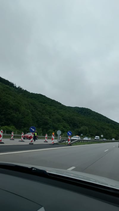 The image depicts a highway scene in Dolna Vasilitsa, Bulgaria, on a cloudy day. A red car and a person in a high-visibility vest are stopped on the side of the road, surrounded by orange and white traffic cones and signs indicating a detour. Further down the highway, several other vehicles are visible, including vans and cars. The background is dominated by a lush, green, tree-covered mountain slope. The perspective is from inside a vehicle, looking out over the dashboard.