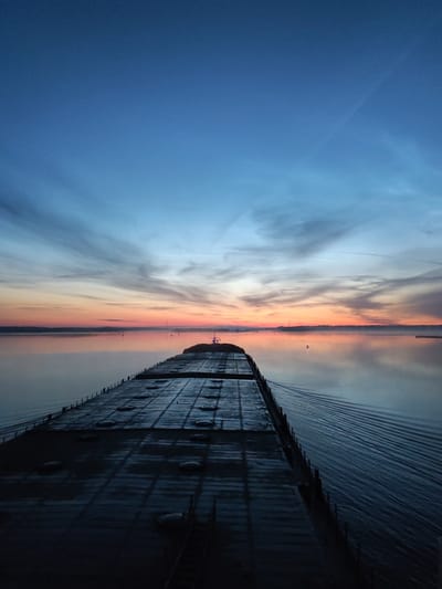 An aerial, long-shot photograph captures a cargo ship sailing on a calm body of water at dawn. The sky transitions from a deep blue at the top to a soft orange and pink hue near the horizon, with wispy clouds stretching across the expanse. The surface of the water is smooth, reflecting the colors of the sky.
The ship, a large, flat-topped vessel, dominates the foreground, extending into the distance. Its deck is dark and appears to be made of metal, with a grid-like pattern of hatches or covers. Faint ripples emanate from the stern of the ship, indicating its movement.
In the distance, a faint silhouette of a landmass is visible along the horizon, suggesting the presence of a coastline or islands. A few buoys or markers are scattered across the water. The scene conveys a sense of peacefulness and solitude, characteristic of early morning on the water. The location is identified as the city of Popovo, Russia.