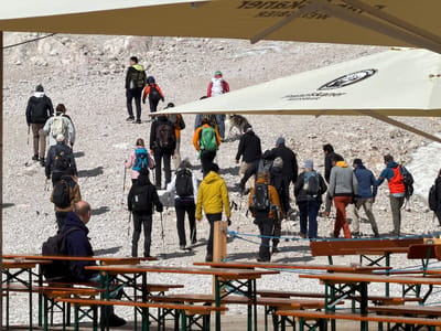 A group of hikers are walking up a rocky, mountainous terrain. In the foreground, several wooden picnic tables are set up under large umbrellas. The umbrellas are cream-colored with logos on them. One umbrella has the logo "Franziskaner Weissbier" and another appears to have some text in a foreign language. The hikers are dressed in outdoor clothing, and many are carrying backpacks and using trekking poles. Some of the hikers are walking together, while others are spread out. A dog is visible walking with some of the hikers. The environment is bright and sunny. The scene appears to be a recreational area in the mountains, possibly a restaurant or rest stop, located in or near Wildermieming, Austria.