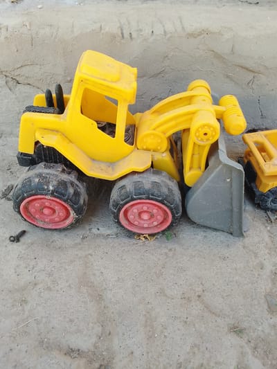 A yellow toy construction vehicle with a bucket loader is positioned on a dirt or sand surface. The vehicle has large, black tires with red rims and appears to be dirty. Behind the loader, a yellow toy dump truck is partially visible. The background consists of a rough, textured wall. The scene is outdoors and appears to be in a somewhat dusty or unkempt area. The image was taken in Muborak, Uzbekistan.