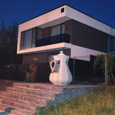 The image shows a modern, two-story house with a dark gray facade and white trim. A large, white, teapot-shaped sculpture is positioned on a set of stone stairs leading up to the house. The sculpture has a window-like opening with stained glass. A dark fence encloses the property, and there is overgrown grass and vegetation around the base of the house. The sky is a deep blue, indicating it is nighttime or twilight. The image appears to be taken in Tohatin, Moldova.