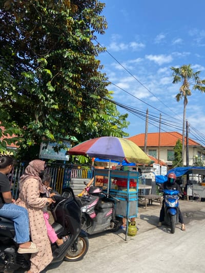 The image shows a street scene in Kecamatan Tulangan, Indonesia, located at coordinates -7.482 latitude, 112.648 longitude. A woman in a brown dress is seated on a black motorcycle, with a young girl standing behind her on the motorcycle's footrest. Both are looking to their right at an unidentified person off-camera. The woman is wearing a headscarf. Another woman is seated on a second motorcycle behind the first motorcycle and is looking towards the camera. A man is on a blue motorcycle parked to the right of the image. He is wearing a black helmet and a blue and white jacket. In the background, there are two buildings, one with a red roof and the other with a green roof. A colorful umbrella with a stand is located in the middle of the image, next to a street food cart. Several power lines are visible overhead. The image suggests a normal day in a small village, with people going about their daily business.