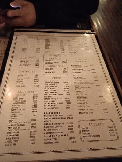 A wine and cocktail menu is displayed on a table in a dimly lit restaurant in Victoria, Argentina, located at latitude: -34.452, longitude: -58.54. The menu lists a variety of cocktails, including classics such as the Old Fashioned and the Margarita. The menu also includes a selection of wines, beers, and spirits. The restaurant has a rustic feel and is dimly lit with soft lighting. The table is wood with a brown finish. The menu is printed on white paper.  The menu items are all written in Spanish.  The prices are listed in Argentine pesos.  There are no people visible in the image.