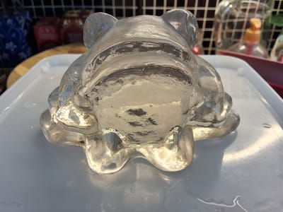 The image shows an ice sculpture of a frog resting on a white plastic tray. The sculpture is transparent and appears to be melting, with water pooling around its base. The background is blurred, but suggests an indoor environment with shelves and various objects, including what appear to be jars or bottles. There are no people or animals visible in the image. The lighting is bright, causing reflections on the ice. The overall scene is static and focused on the ice sculpture. The provided information about Arboga, Sweden does not appear to be relevant to the visual content of the image itself.