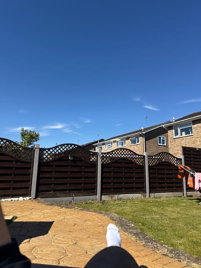 The photograph depicts a suburban scene in Colchester, United Kingdom. A clear blue sky with wispy cirrus clouds dominates the left side of the frame. On the right, a residential property is visible, featuring a two-story brick building with white-framed windows. A dark brown wooden fence with a decorative lattice top runs along the property line, separating it from a green lawn. Clothes hang on the fence. The shadow of a person wearing a white sock is visible in the bottom right corner. The ground is covered with textured, yellow paving stones.