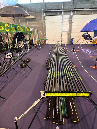 The image displays an indoor exhibition or sales event for fishing equipment, located in Mora, Portugal. The venue features a blue carpeted floor, light-colored concrete walls, and two large white industrial roll-up doors in the background. Overhead lighting fixtures are visible.

In the foreground and extending into the midground, a substantial quantity of fishing pole sections, predominantly black with green and yellow "SENSAS" branding, are laid out horizontally on black tripod display stands. The foremost display features yellow price tags showing various figures, including 2399, 1999, 1550, 899, 1918, and 239.99. Beneath this display, several black and yellow "MAMMOTH" branded rod cases and additional pole sections are visible on the floor.

To the left, a "Sensas World Champion" branded black umbrella is open over a booth. A green banner with "Sensas" branding marks this area. One individual, partially obscured by the display, is visible within the booth. Additional fishing rods are arranged on stands in this section.

To the right, a blue umbrella covers another display area. A white and red barrier rope, supported by white and red bollards, delineates a pathway. A blue mat with "CC CUMIC" is on the floor beyond the barrier. A blue industrial trash bin is situated near the rightmost roll-up door.