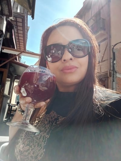 A woman is depicted in a selfie-style close-up, holding a glass of red liquid outdoors under bright sunlight in El Espinar, Spain. The subject has long, dark brown hair and wears large, dark sunglasses that obscure her eyes. Her lips are painted with pink lipstick. She is wearing a dark, possibly black, top adorned with a pattern of shiny studs or beads on the chest. On her left wrist, she wears a multi-strand bracelet.
Her left hand holds a large, stemmed glass filled with a dark red beverage, likely wine or sangria. Reflections within the sunglasses and the glass reveal an outdoor setting, including at least one other person seated opposite the subject, along with tables and chairs, indicating a cafe or restaurant terrace.
The background shows a clear blue sky with bright sunlight causing lens flares in the upper right quadrant. To the left, a building with wooden structures, possibly awnings or balconies with metal railings, is visible. To the right, another building constructed of exposed red brick features balconies. The scene suggests an outdoor dining or social area within the town of El Espinar, Spain; however, a precise street address or specific landmark is not discernible from the image.