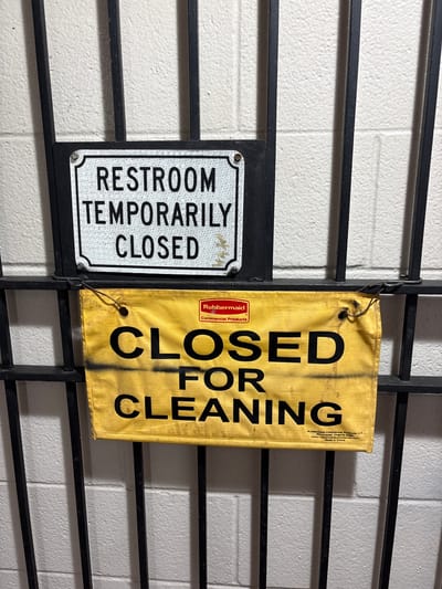 A close-up, eye-level shot shows two signs attached to a black metal gate in Sonora, California, United States. The gate is comprised of vertical bars. Behind the gate, a white cinder block wall is visible.
The upper sign is a white, rectangular plaque with a silver reflective border. It is attached to the gate with a screw at the top left corner. The text on the sign reads, in black sans-serif font, "RESTROOM TEMPORARILY CLOSED".
The lower sign is a yellow, rectangular banner with black text and a small red Rubbermaid logo in the top right corner. This sign is attached to the gate using thin wire. The banner is wrinkled and appears to be made of a fabric material. The black text on the banner is larger and bolder than the text on the upper sign. It reads, in three lines: "CLOSED FOR CLEANING". Beneath the word "CLEANING," smaller text indicates "Rubbermaid Commercial Products LLC, Winchester, Virginia 22601, www.rubbermaidcommercial.com, Made in China." The banner also has some smudges of black spray paint or dirt.