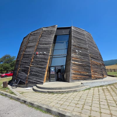 In Dorkovo, Bulgaria, a distinctive, conical wooden building stands against a clear blue sky. The structure is clad in dark, weathered horizontal wooden planks, with some showing a reddish-brown undertone. A large, angled glass facade is set into the front of the building, revealing an entrance with glass doors. Two individuals are visible standing just inside the entrance. To the left of the building, a bright red car is parked on a grassy area next to a tree. A small table with two chairs is also situated near the building. The ground in front of the entrance is paved with square concrete tiles, and a concrete walkway leads to the building's entrance. In the background, a grassy hillside slopes upward, with a wooden fence visible along its crest. The scene is bathed in bright sunlight.