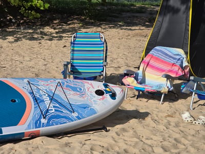In the city of Flowery Branch, United States of America, a sandy beach setting is depicted, bathed in bright sunlight. In the foreground, a colorful paddleboard with blue, white, and pink patterns rests on the sand, with a paddle lying nearby. Behind the paddleboard, two striped beach chairs are set up. One chair, predominantly blue and green, is empty, while the other, draped with a striped towel in shades of pink, blue, and yellow, is also unoccupied. To the right, a black and yellow portable shelter stands partially open. Further to the right, a pair of white clogs with floral patterns are placed on the sand. The environment is natural, with sand, shadows from trees, and distant greenery suggesting a waterside location.