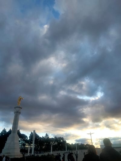 The image depicts an outdoor scene within the Sanctuary of Our Lady of Fátima, Portugal, specifically the Esplanade of the Sanctuary. The upper two-thirds of the frame are dominated by an overcast sky with dense, dark grey clouds interspersed with patches of lighter grey and a small area of blue sky in the upper left. A band of brighter, illuminated clouds is visible along the horizon, suggesting dawn or dusk.
In the lower left quadrant, a tall, white stone column rises, topped with a golden statue, likely representing the Sacred Heart of Jesus. The figure faces right, with its right arm extended and hand pointing outwards. To the right, in the mid-ground, a large, dark-colored, modern crucifix is silhouetted against the brighter horizon.
The ground level features a vast, open paved area, the Esplanade, populated by numerous small, indistinct figures of people scattered across the space, appearing to walk or stand. A line of dark green trees forms a boundary behind the Esplanade, partially obscuring distant white buildings and several tall, slender white columns. In the immediate lower right foreground, the backs of two individuals are partially visible, facing away from the camera and towards the scene.