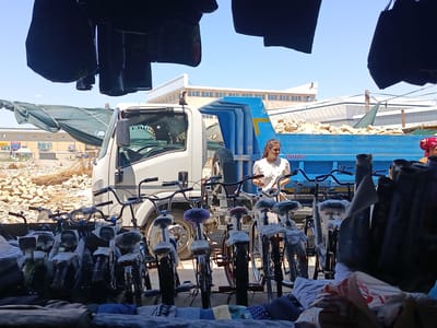A young woman wearing a white top is standing next to a blue dump truck in a sun-drenched outdoor market in Alat, Uzbekistan. The dump truck is filled with light brown rocks and debris. In front of the truck, several bicycles are lined up, some covered in plastic. The foreground of the image shows more bicycles and stacks of fabric. The background reveals a long, light-colored building with many windows and a clear blue sky above. The scene suggests a commercial setting, possibly a sale or a delivery.