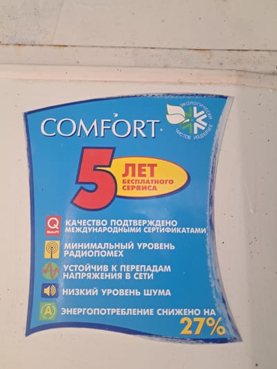 The image displays a close-up view of a blue label with white and yellow text. The label features a large red number "5" followed by the word "ЛЕТ" (years) in yellow. Below this, the text "БЕСПЛАТНОГО СЕРВИСА" (free service) is also in yellow.

Several icons and text lines are arranged in a list format on the blue background. These include:
- A red "Q" with "QUALITY" written below it, followed by text related to international certifications.
- An icon of a radio tower, with text indicating minimal radio interference.
- A wavy line icon, with text stating stability against voltage fluctuations.
- A speaker icon, with text indicating low noise levels.
- A green circular emblem with "A" inside and Russian text, followed by text about reduced energy consumption by 27%.

The label itself is glossy and appears to be affixed to a white surface. The white surface has some minor blemishes and what appear to be specks of dirt or dust. The top of the image shows a horizontal white surface with some rust-colored streaks.

The provided context mentions the location as Chobruchi, Moldova. However, there are no visual cues within the image itself that directly indicate this specific geographical location. The image is a product label and does not contain any elements that would allow for its localization to Chobruchi or any other city.