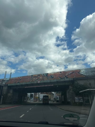 The image depicts a view from inside a vehicle moving along a road under a railway bridge in Osnabrück, Germany. The sky, predominantly filled with white and grey clouds, is visible on the left. The railway bridge, marked with graffiti, runs horizontally across the upper portion of the frame. A street runs under the bridge with a vehicle partially visible. Signage is present along the side of the road and under the bridge. The road appears to be asphalt, with red lines demarcating the lane boundaries. A green sticker is visible in the foreground on the right, possibly attached to the vehicle’s window.