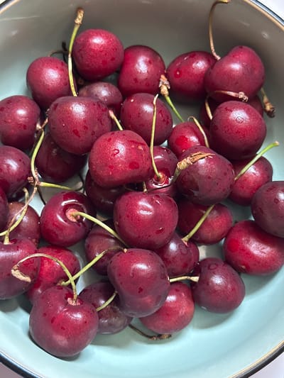A close-up shot shows a bowl filled with dark red cherries with green stems. The cherries appear fresh, some with water droplets on their surfaces. The bowl has a pale blue interior and a dark rim. The scene is illuminated, highlighting the textures and colors of the fruit. No specific location in Herxheim, Germany, can be determined from the image. No people are visible.