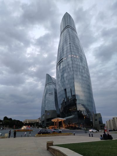 This is a low-angle, eye-level, outdoor, full shot photograph of the Flame Towers in Sabail Raion, Azerbaijan. The towers are two skyscrapers that resemble flames and are covered in glass. The sky is cloudy and gray. In the foreground, there is a paved walkway and a grassy area. Several people are visible on the walkway and in the distance, and there are cars on the road.
