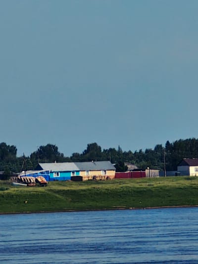 The image depicts a waterfront scene in the village of Potapovo, Russia. In the foreground, a wide body of blue water with rippling surface is visible. The middle ground features a grassy embankment rising from the water, topped by a line of buildings and vegetation. The buildings appear to be houses, with roofs of various colors including silver, blue, and yellow. Some houses have visible windows and doors. To the left of the buildings, a small boat or barge is partially visible on the grassy area, near some brown, rusted metal objects. Beyond the houses, a dense line of dark green trees forms a backdrop. In the sky above, a pale blue expanse stretches across the upper portion of the image. The overall atmosphere is serene and suggests a rural or semi-rural setting.
