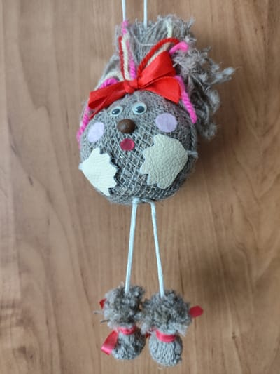 A handmade toy hangs against a wooden surface. The toy's head is a burlap-covered sphere adorned with googly eyes, a brown button nose, pink felt cheeks, a red felt mouth, two cream-colored patches on the lower face, a red ribbon, and yarn strands. Two white strings hang from the head, each ending in a burlap-covered ball with a red ribbon. The background is a wooden surface with visible wood grain. The location is Iwye, Belarus.