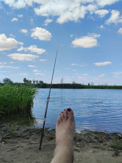 This media file depicts a serene outdoor scene of fishing by a lake under a bright, cloudy sky. The foreground is dominated by a bare foot with dark nail polish, suggesting a relaxed and casual atmosphere. To the left, lush green reeds grow by the muddy shoreline, indicating a natural, perhaps marshy, environment. A fishing rod stands upright in the sand, its line cast into the calm, blue water.
The middle ground showcases the expansive lake, its surface rippled by a gentle breeze. The far shore is lined with dense vegetation and scattered trees, some appearing bare or dormant, suggesting a temperate climate or a specific season. The sky above is a vibrant blue, dotted with fluffy white clouds, implying pleasant weather and possibly midday.
The overall mood is one of tranquility and solitude, ideal for a quiet fishing trip. There are no visible text elements in the image. The combination of natural elements and the fishing activity points to a peaceful recreational outing in a lakeside setting. While the prompt provides "San Martín de Porres, Peru" as location context, the visual elements themselves do not offer specific cues to confirm or deny this location, appearing more generically as a body of water with surrounding vegetation.
