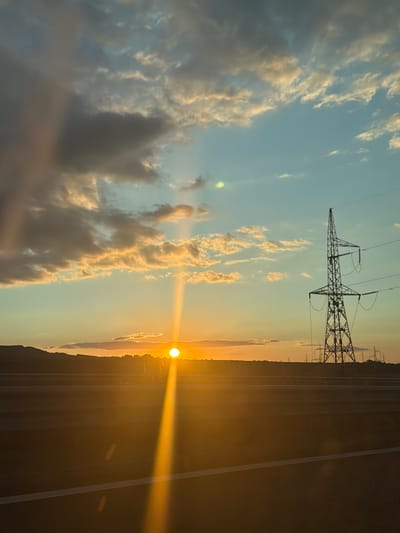 This is a vertical, medium shot of a sunset over a highway in or near Studena, Bulgaria. The sun is a bright, glowing orb low on the horizon, casting a warm, golden light across the scene. A distinct sunbeam extends diagonally upward from the sun. The sky is a gradient of pale blue at the top, transitioning to hues of orange and yellow near the horizon. There are scattered clouds, some dark and gray, others tinged with the warm light of the sunset.
In the middle ground, a dark silhouette of a landscape with hills and vegetation stretches across the frame. A prominent electricity pylon with a lattice structure stands tall on the right side of the image. Several other, smaller electricity pylons are visible in the distance to the right, suggesting a power line corridor. The foreground is dominated by the dark surface of a highway, with a white line marking the lane. The bright sunlight creates lens flares and reflections on the highway surface, obscuring details. The scene is devoid of people. The overall atmosphere is serene and captures the natural beauty of a sunset combined with man-made infrastructure.