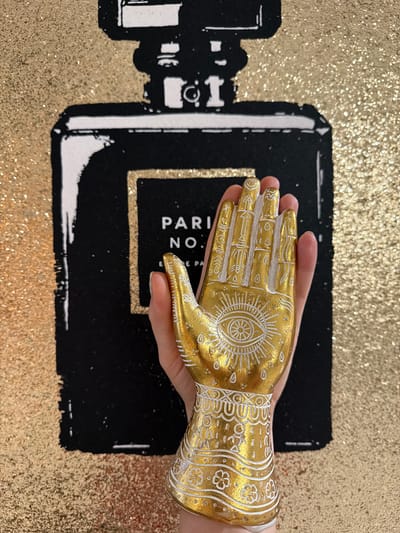 A hand holds a golden, decorative hand-shaped object in front of a black and gold sparkly backdrop featuring a stylized perfume bottle. The backdrop is a painting or print of a black perfume bottle with white accents on a gold glitter background. The perfume bottle has "PARIS NO." and "NE PA" printed on it in white. The golden hand-shaped object is decorated with white patterns, including an eye symbol on the palm and floral designs on the wrist. The background texture suggests sequins or glitter, creating a shimmering effect. The scene is indoors, with even lighting. No specific location in Riverview, United States of America, is discernible from the image.