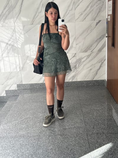 A young adult female is depicted standing in an indoor setting, taking a mirror selfie with a white smartphone held in her right hand. She has dark, shoulder-length hair and a neutral facial expression. She is dressed in a dark green, patterned, strappy mini-dress with a sheer hemline, dark brown ankle socks with reversed white text, and olive green sneakers with white soles. A black shoulder bag with a silver chain accent is carried on her left shoulder. The background features a wall composed of rectangular white marble tiles with grey veining. The floor is covered with large, speckled grey granite-like tiles. A partial view of a matching grey tiled staircase is visible on the left. On the right, a light brown wooden door or panel is present, along with a mounted rectangular unit above it. The scene is illuminated by indoor lighting, casting a faint rectangular shadow on the lower right portion of the floor. This scene is located in Ciudad del Libertador General San Martín, Argentina.