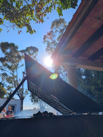 A tilted, low-angle shot captures a sunny outdoor scene. A dark, gridded grill occupies the right side of the frame. In the background, a white building and greenery are visible under a clear blue sky. The sun shines brightly, creating lens flare. The scene suggests a summer day, potentially a barbeque, with the grill as the focal point.