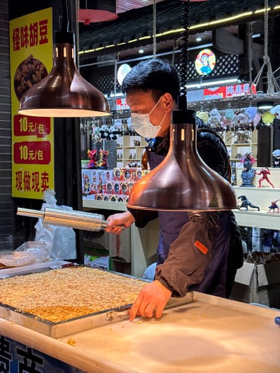 An adult male is observed working at a food stall in Pengshui Miao and Tujia Autonomous County, China. He wears a light blue surgical mask, a dark blue apron, and a dark long-sleeved garment. Two copper-toned, bell-shaped heat lamps are suspended overhead, illuminating the workspace. The man's right hand holds a textured silver metal roller, while his left hand points at or touches a few kernels on a flat, white work surface. To his left, a rectangular metal tray contains a substantial quantity of small, yellowish-brown, granular food, consistent with roasted broad beans. A yellow sign on the far left advertises "怪味胡豆" (Strange Flavor Broad Beans) for "10元/包" (10 yuan/bag) and states "现做现卖" (Made fresh, sold fresh). The background features a retail display with various items, including plush panda figures, boxed merchandise, and superhero figurines such as Spider-Man and Batman. Decorative elements include a red paper umbrella and a circular illuminated sign depicting a traditional Chinese opera character. The stall's structure includes dark brickwork and an overhead dark roof, indicating an outdoor or market setting.