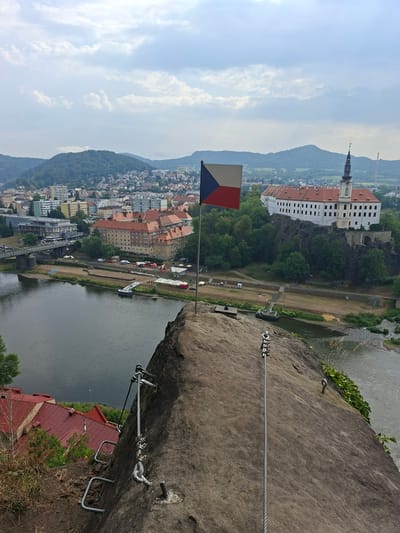 An eye-level, high-angle outdoor shot captures a view of Děčín, Czechia, from a rocky outcrop. In the foreground, a Czech flag flies from a flagpole on the rock. Metal rungs and a cable are visible on the rock face, suggesting a climbing or via ferrata route.

Below, the Elbe River flows, with a sandy bank and several tents set up on it. A small boat or dock is visible in the river. Across the river, a bridge connects to the city, which features a mix of modern and older buildings, including a prominent white building with a red roof and a tower on a rocky hill. Distant green hills and mountains form the backdrop under a cloudy sky. The scene appears to be a scenic overlook or a point of interest for tourists.