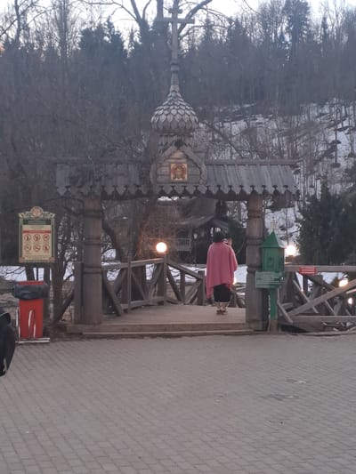 A pedestrian walks across a wooden bridge in Vzglyadnevo, Russia. A traditionally styled wooden gate with an onion dome and religious icon stands at the bridge's entrance. A park information sign and trash bin are positioned nearby. Bare trees surround the structure, and the pedestrian path is paved with bricks.