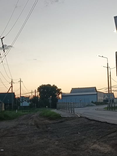 The image shows a rural road with power lines and a few buildings under a pale sky, possibly at dusk or dawn. In the foreground, the road is unpaved and rough. To the left, there are power poles with wires stretching across the frame. A low building with a dark roof is visible on the left. In the mid-ground, a large, light-blue industrial-style building with a corrugated roof is prominent. A bridge with metal railings crosses a road in front of this building. A tree with dense foliage is located to the right of the large building. Streetlights are present on poles along the road. The overall scene suggests a quiet, somewhat industrial or agricultural area on the outskirts of a settlement in Tatarstan, Russia. No people are visible in the image.