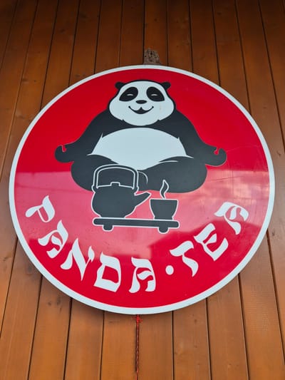 This media file features a circular, red sign with a white border, mounted on a wooden wall. The sign depicts a black and white panda in a stylized, cartoonish manner, positioned upside down and to the left of the sign's center. The panda appears to be wearing a backpack and holding an axe. White lettering, in a decorative, Gothic-style font, arcs around the right side of the panda, appearing to spell out words.
The background is a wooden wall composed of horizontal planks, stained a warm, brown hue. The lighting suggests it is daytime, with no distinct shadows indicating a specific time of day. The environment appears to be outdoors, given the wooden wall and the general aesthetic. There is a thin red string or wire visible on the right side of the image, attached to the sign and extending towards the wooden wall. The sign itself shows some signs of wear, with faint scuff marks visible on its surface. The visible text appears to read "PANDA BEER".