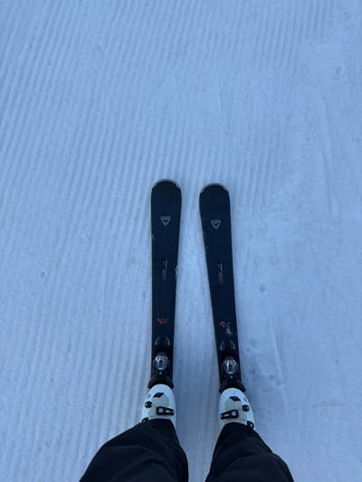 The image depicts a first-person, downward perspective of a person's lower extremities equipped for skiing on a groomed slope in Saanen, Switzerland. Two black Rossignol skis, marked with "LCT" and "N14" in red text, are parallel on the snow. Black and silver ski bindings attach the skis to two white ski boots. The lower portions of black ski pants are visible, covering the top of the boots. The snow surface exhibits distinct, parallel corduroy patterns, indicating recent grooming. No other individuals or structures are present in the visible frame, suggesting a solitary moment on a prepared ski run.