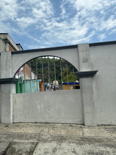 The image presents a close-up view of a concrete structure with an arched opening containing metal bars. The structure appears to be part of a building, possibly a cemetery entrance, as suggested by the view through the bars showing graves and a variety of structures in a town in the city of Jilotepec, Mexico. The sky is visible through the upper portion of the arch, exhibiting a light blue color with scattered white clouds. The concrete of the structure shows signs of aging and weathering. The buildings in the background are low-rise, with a mix of colors and materials.