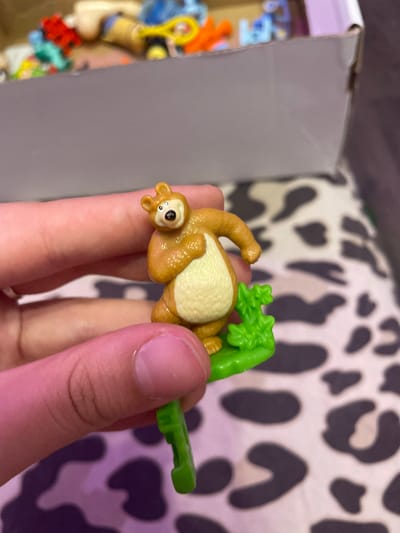 A close-up shot focuses on a small, brown, plastic bear figurine held between two fingers. The bear is standing on a green plastic leaf-like base. Its body is rounded, with a lighter colored belly. It has small ears, a black nose, and open eyes with black pupils. The background is out of focus, showing a cardboard box filled with various small, colorful toys, and a patterned surface that appears to be fabric. The lighting is bright, casting soft shadows. There is no indication of the location being Borisova Griva Massiv, Russia.