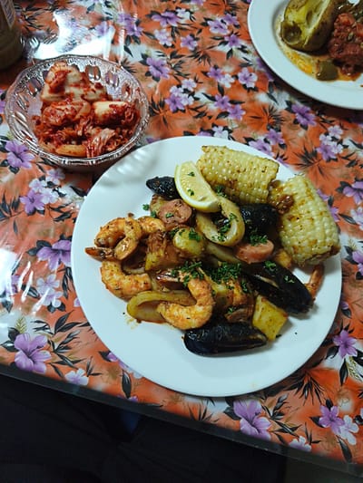 The image displays a close-up, overhead view of a meal served on a white plate, placed on a table covered with an orange floral tablecloth. The meal appears to be a seafood boil, featuring shrimp, mussels, corn on the cob cut into segments, and what seems to be calamari rings, all mixed with pieces of potato and sausage. Garnishes of chopped parsley are visible. To the left of the plate, a small glass bowl contains kimchi, a staple Korean fermented dish. The lighting is natural, suggesting an indoor setting. There is no indication of the specific location in Yeosu-si, South Korea, beyond the presence of food commonly found in Korean cuisine. The scene is static, focusing solely on the presented food.