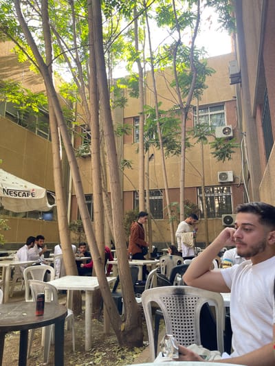 The image depicts an outdoor cafe or courtyard scene in قرية الضبية, Syria, during daylight hours under bright sunlight. The foreground features a young man with dark hair and a beard, wearing a white t-shirt, seated at a white plastic chair on the right, looking to the right with his right hand resting on his chin. A silver foil object is on the table in front of him. To his left, a dark round table holds a red and black beverage can.
In the midground, multiple white plastic square and round tables with white plastic chairs are occupied by several individuals. On the left, three young men are seated at a white table; two wear white thobes and appear to be conversing. Further right, a young man in a brown jacket stands near a table, and another in a light-colored long-sleeved shirt stands holding a white cloth. Approximately 10-15 individuals, primarily young adults, are visible throughout the seating area.
Numerous slender, tall trees with light green foliage punctuate the scene, casting shadows and dappled light. The ground consists of uneven dirt or loose earth. In the background, beige-colored multi-story buildings with multiple windows and several external air conditioning units are visible. A white parasol with a "NESCAFE" logo is open on the far left. A darker building section is partially visible on the far right.
