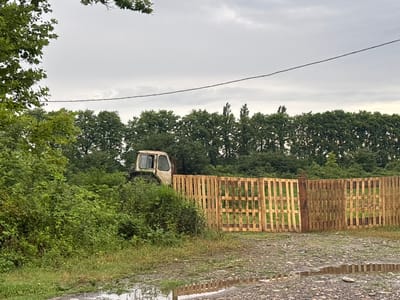 The image depicts an outdoor scene in the village of Nigotisa, Georgia. A white, weathered tractor is partially obscured by lush green foliage on the left side of the frame. To the right of the tractor, a newly constructed fence made of light-colored wooden pallets stretches across the scene. The fence has a gate-like section with vertical planks. The ground in the foreground is a mix of gravel and muddy puddles, with reflections visible in the water. A dense line of green trees forms a backdrop behind the fence and foliage. The sky above is overcast and gray, with a thin power line stretching horizontally across the upper portion of the image. No people are visible in the frame.
