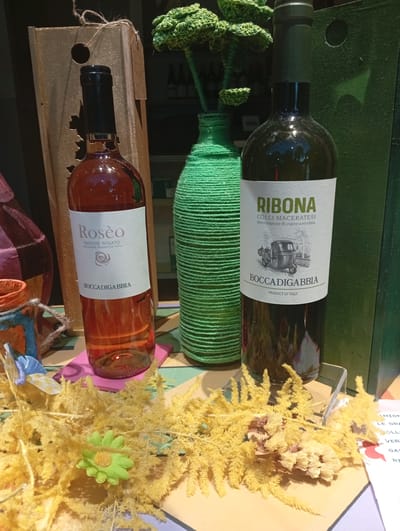The image shows a retail display featuring two bottles of wine and decorative elements. On the left is a bottle of "Rosè" wine with a white label that reads "MARCHE ROSATO" and "BOCCADIGABBIA." To its right is a bottle of "RIBOBA" wine, also with a white label, displaying "COLLI MACERATESI" and "BOCCADIGABBIA PRODUCT OF ITALY," and an illustration of a three-wheeled vehicle. Between the bottles is a tall, green, textured vase. In the foreground, there are arrangements of dried yellow foliage and a small green artificial flower. A wooden display stand with a cutout is behind the rosé bottle, and a dark green display stand is behind the Riboba bottle. The scene appears to be indoors, likely a shop or market stall, possibly in Civitanova Marche, Italy, given the presence of Italian wine labels.