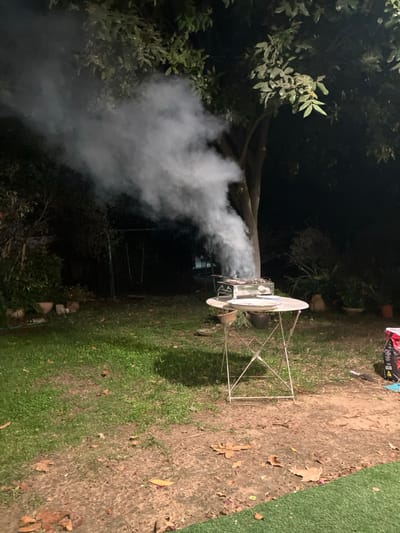 The image displays an outdoor scene at night in the Brener Regional Council, Israel. In the midground, a white, round metal folding table supports a rectangular charcoal barbecue grill. The grill is actively producing a significant, dense plume of gray smoke that ascends vertically and then drifts towards the upper left. A white plate or tray is visible on the table next to the grill. On the ground to the right of the table, a red and black bag, appearing to be charcoal, is present. The ground consists of green grass mixed with patches of exposed soil and scattered dry leaves. A section of artificial green turf is visible in the lower right foreground. In the background, a large tree with leafy branches is prominent on the right, and additional dense foliage and smaller trees are on the left. A faint chain-link fence and a swing set are discernible further back in the darkened central area. Several potted plants are arranged along the left side of the scene. The area is illuminated by artificial light, creating distinct shadows and highlighting the smoke and foreground objects. No people are visible.