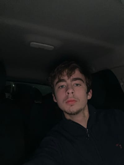 A person with fair skin, dark hair, and a light beard looks into the camera. The person is wearing a dark-colored, zippered jacket. The person is positioned in what seems to be the interior of a car; a grey side panel of a car is visible in the background, along with a car's interior light panel and side mirror. The lighting in the image is dim.