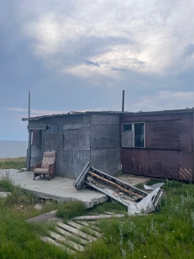 The image depicts an abandoned, dilapidated structure on a coastal landscape. The building appears to be made of various salvaged materials, including weathered plywood and corrugated metal. A single upholstered armchair sits on a concrete slab in front of the structure. To the right, a large, broken concrete slab rests precariously, with what appears to be debris or a section of a metallic structure on top of it. The surrounding environment is overgrown with green grass and sparse vegetation. The sky above is overcast with dramatic clouds. The distant horizon shows a body of water, likely the sea or a large gulf. The overall scene evokes a sense of desolation and decay. The specific location within Yamburg, Russia, is not explicitly identifiable from the visual information.